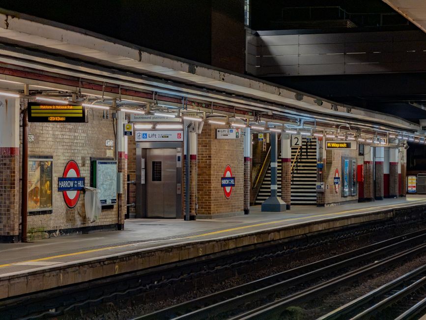 A view of Hornchurch underground station platform during nighttime, featuring brick walls and signage indicating the station name as 'Harrow & Hillingdon.' The platform has a tiled floor with a yellow